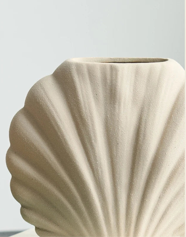 A close-up of a sculptural ceramic vase with a seashell-inspired ribbed design, in a warm cream or off-white glaze, set against a neutral light gray background.