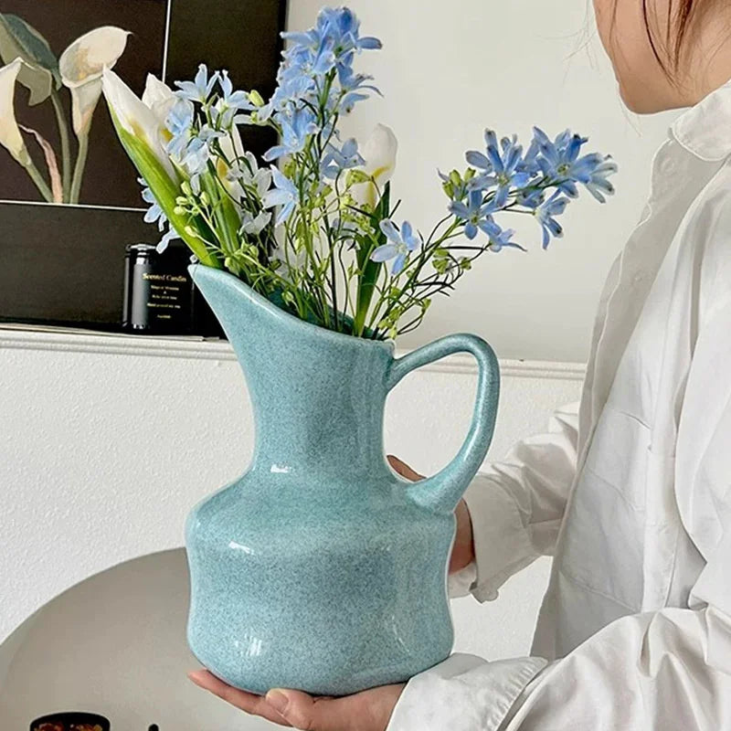 A person in a white shirt holds a speckled light blue ceramic pitcher vase filled with blue flowers and green stems in an indoor setting.