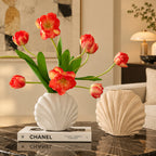 Two sculptural shell-shaped vases displayed on a dark round table. The left vase is white and sits on a white book, holding coral-pink tulips with green leaves.