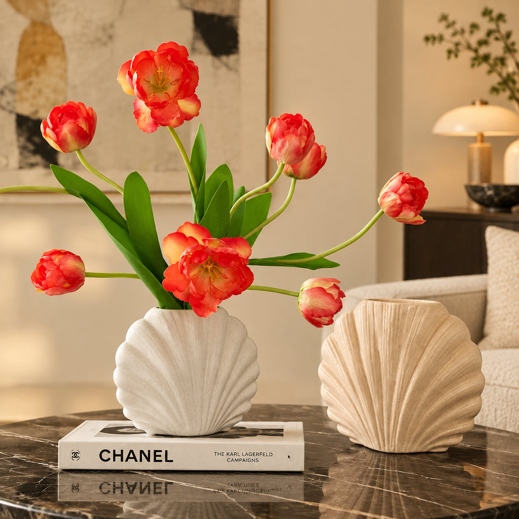 Two sculptural shell-shaped vases displayed on a dark round table. The left vase is white and sits on a white book, holding coral-pink tulips with green leaves.