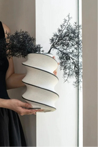 A woman in a black sleeveless dress holds a sculptural, tiered off-white ceramic planter with black rims, which is styled with two black, branching decorative plants against a bright window.