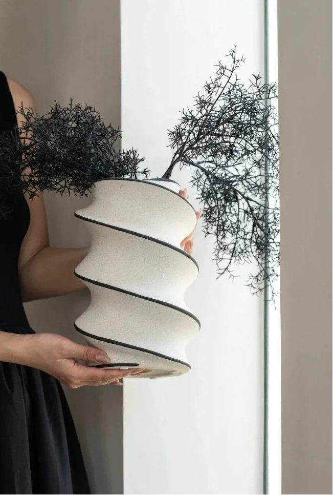 A woman in a black sleeveless dress holds a sculptural, tiered off-white ceramic planter with black rims, which is styled with two black, branching decorative plants against a bright window.
