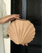 A person holds a light beige, shell-shaped decorative vase or planter with a ribbed texture. The background features a split design with a light beige wall on the left and a dark black door on the right.