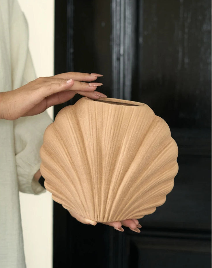 A person holds a light beige, shell-shaped decorative vase or planter with a ribbed texture. The background features a split design with a light beige wall on the left and a dark black door on the right.