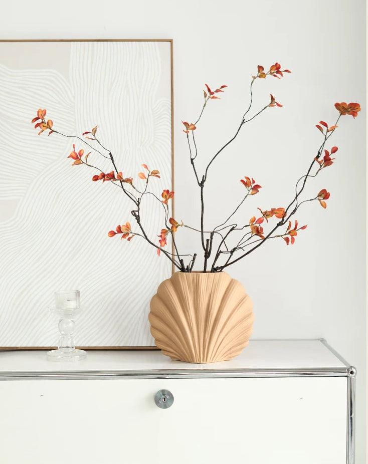 A beige scalloped ceramic vase filled with dried branches and small orange-red leaves, displayed on a white cabinet against a light wall with framed wall art.