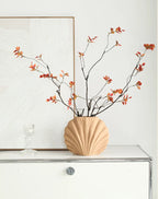 A beige scalloped ceramic vase filled with dried branches and small orange-red leaves, displayed on a white cabinet against a light wall with framed wall art.