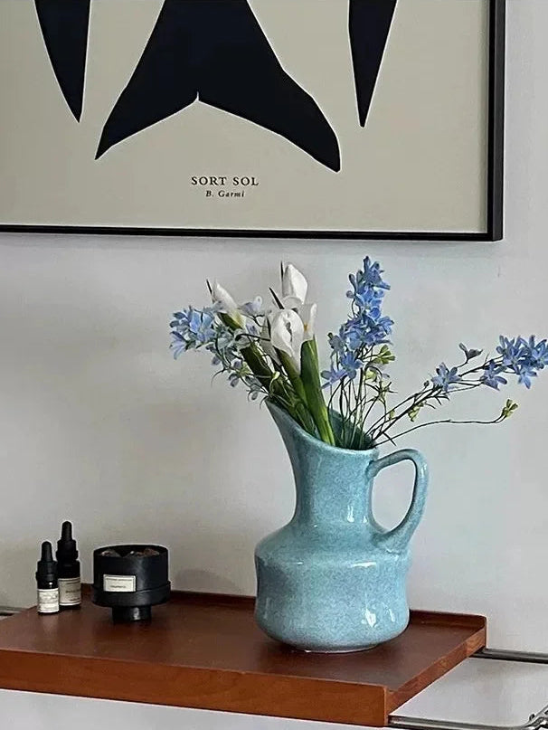 A light blue speckled ceramic pitcher vase filled with white and blue flowers, styled on a wooden floating shelf against a white wall with a framed print.