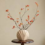 A beige ribbed ceramic vase holding black wire branches with small orange and red leaves, displayed on a dark wooden pedestal against a warm beige wall. The text '@CRUX' appears on the right side.