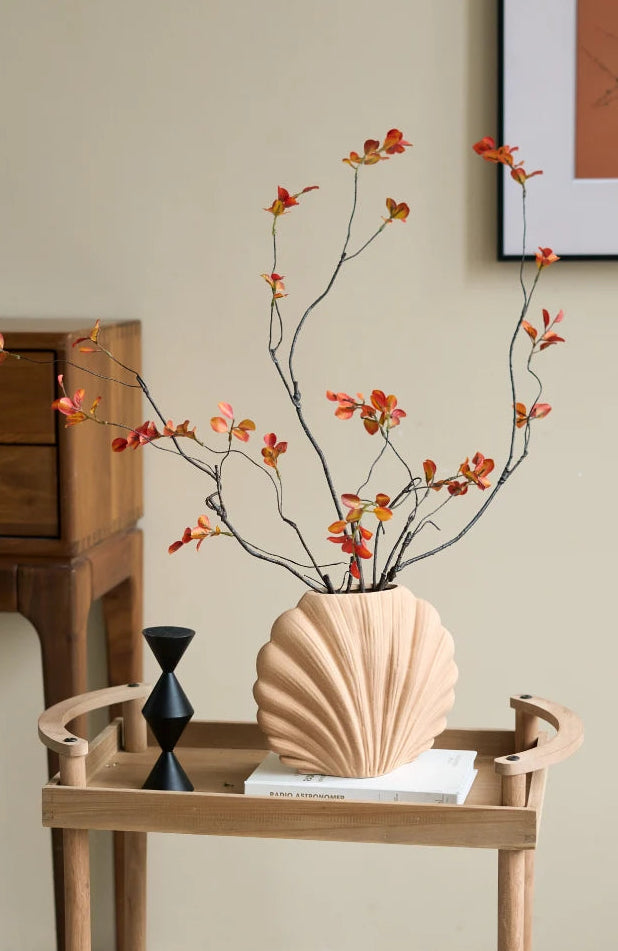 A beige scallop shell-shaped ceramic vase filled with dried branches and small orange-red leaves, displayed on a light wood side table in a styled interior setting.