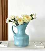 A light blue ceramic pitcher vase filled with white and pale yellow flowers, styled on a white shelf next to two books against a white wall.