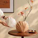 A beige ribbed ceramic vase with a scallop shell shape sits on a round wooden table, holding a dried branch with small orange flowers. A person's hands are shown holding a matching white ribbed shell-shaped object.