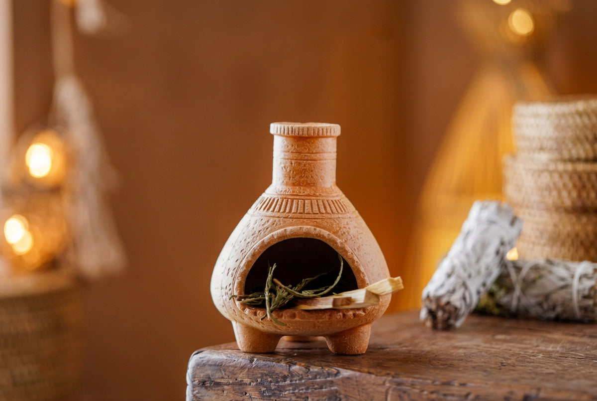 Small terracotta oven with wooden logs and greenery on a wooden surface, with a blurred background.