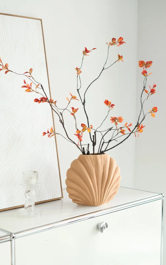 A beige ribbed ceramic vase filled with dried branches and small orange-red flowers, displayed on a white cabinet in a bright interior setting.