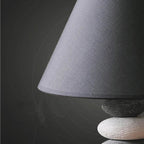 A close-up of a table lamp featuring a large dark gray fabric cone shade and a stacked stone base in dark and light gray.