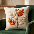 A square cream throw pillow with a 3D embroidered leaf design in orange and beige, resting on a wooden chair. Text at the top reads '2D Flat Printing, Non-Real Embroidery'.