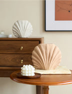 A beige pleated shell-shaped vase displayed on a round wooden table in a styled interior setting, alongside a white sculptural candle and a wooden cabinet in the background.