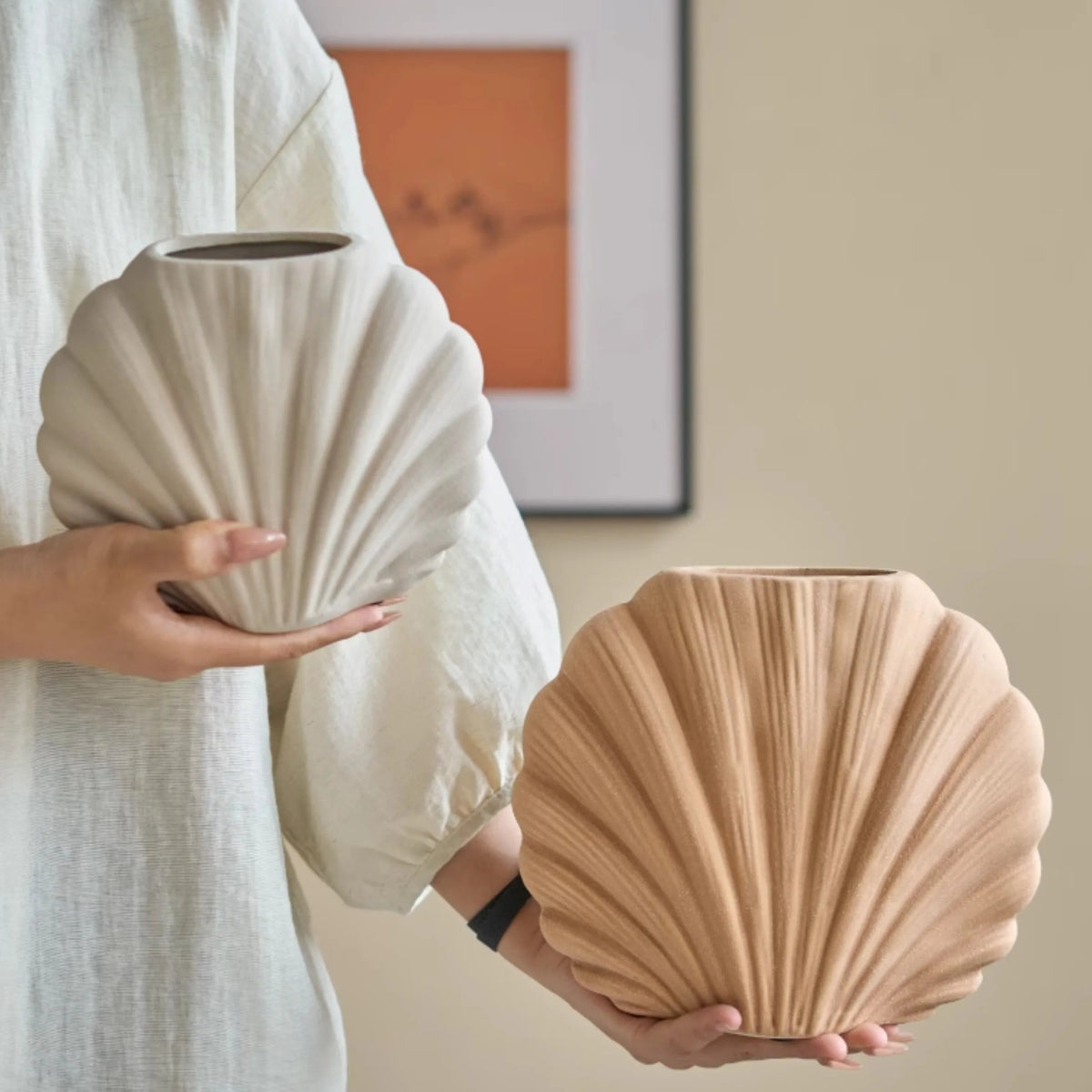A person in a white robe holds two ribbed ceramic vases in a warm neutral interior. The vases are the product, shown in a styled room scene with wall art and a beige wall behind them.