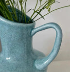 A close-up of a speckled light blue ceramic pitcher vase with a large handle, containing green stems against a plain light background.