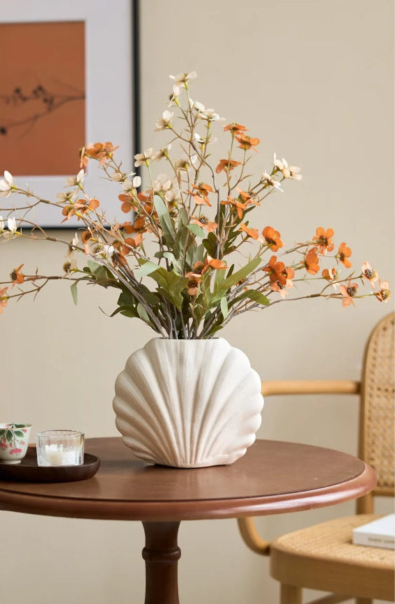 A ribbed white ceramic vase filled with dried orange and white flowers, placed on a round wooden table in a warm, neutral-toned room with a woven chair and framed wall art in the background.